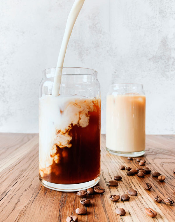 Iced coffee with almond milk being poured in and a glass of almond milk in the background